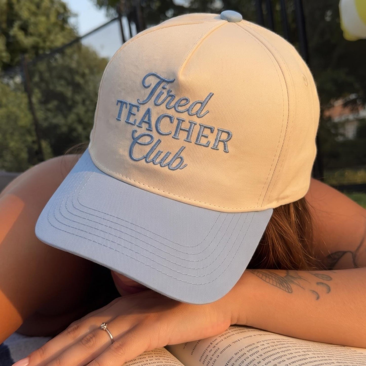Tired Teacher Club' baseball cap in cream and blue with embroidered text, worn by person reading outdoors
