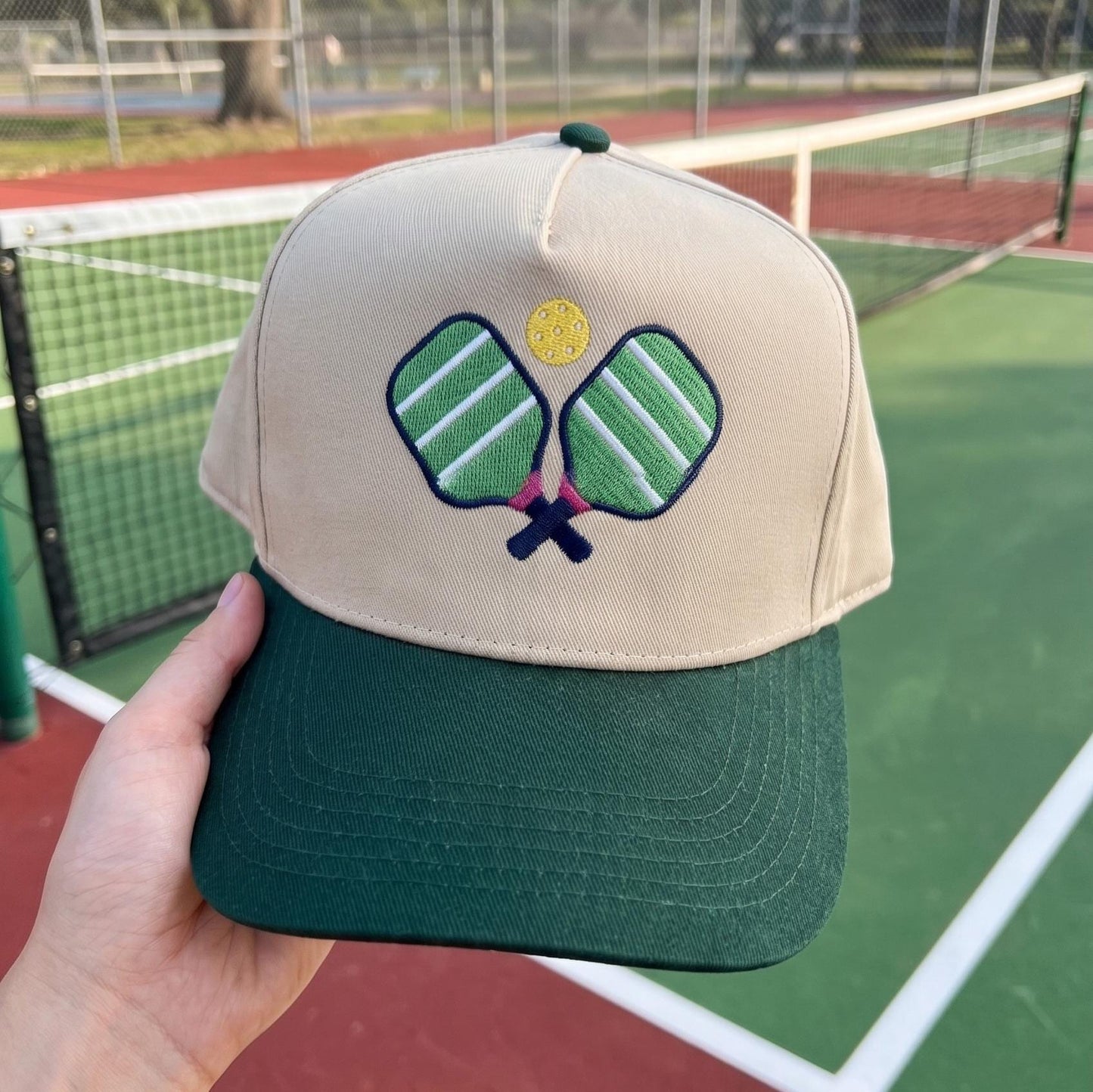 Pickleball cap with embroidered paddles and ball, beige crown with green brim, on a sports court